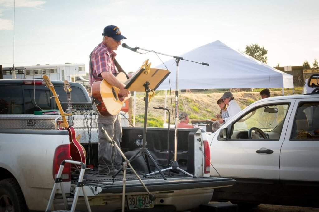 A musician performs on a pickup truck at an outdoor event. A white tent and harvested field are visible in the background.