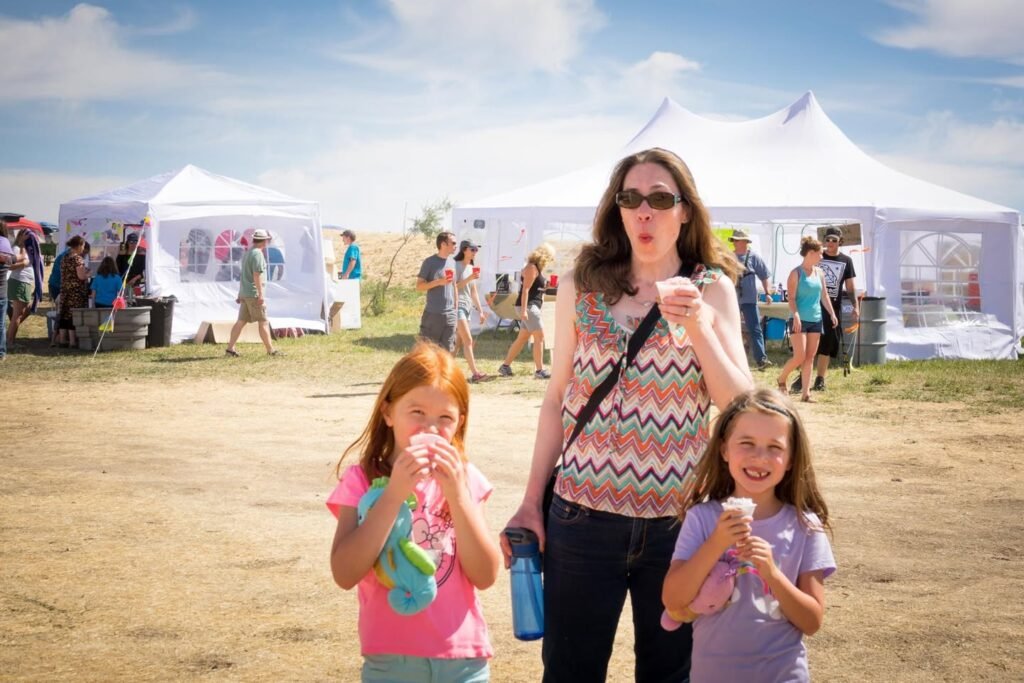 A family enjoys ice cream cones at an outdoor event with white vendor tents under a sunny sky.