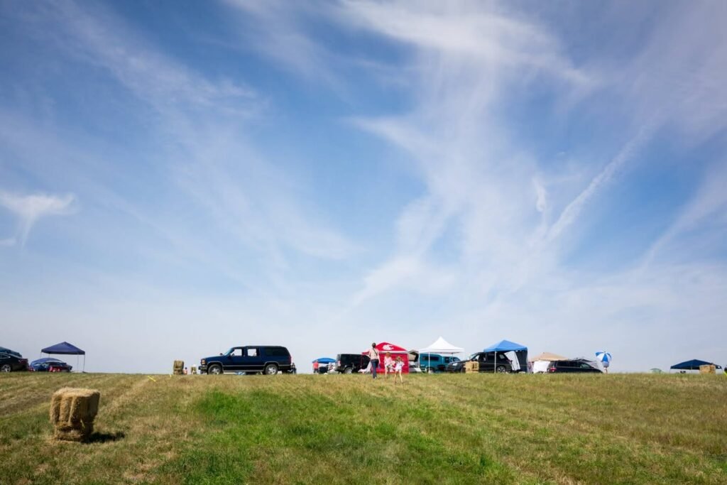 Vehicles and tents are set up on a grassy field under a blue sky. Hay bales and umbrellas are visible around the area.