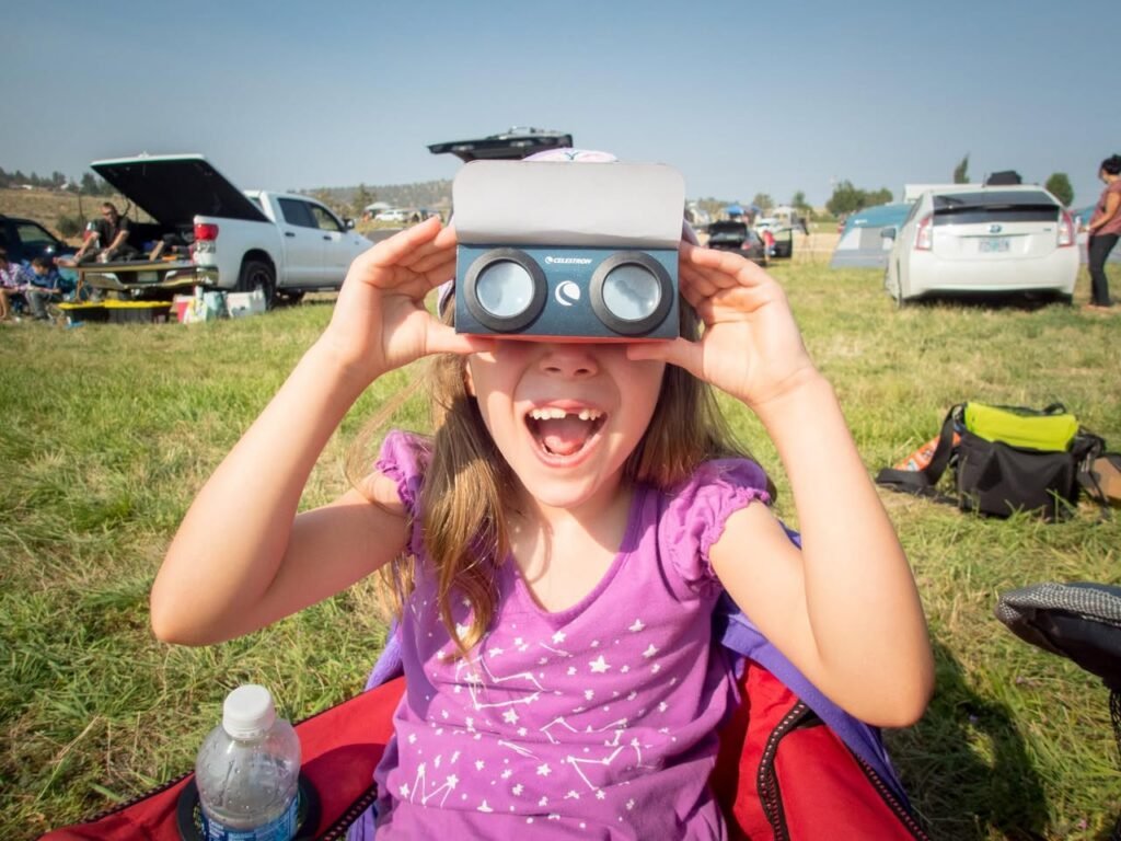 A person holds a diorama viewer to their eyes in a grassy parking area, with cars and outdoor gear in the background.
