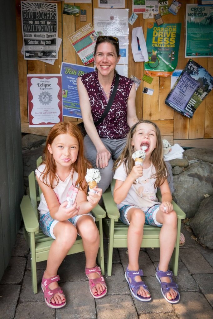 A woman and two children enjoying ice cream cones while seated on small green chairs in front of a poster-covered wooden wall.