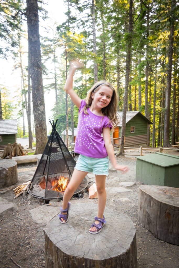 A person in a purple shirt standing on a log stump waving their hand next to a fire pit in a forest setting. Firewood and tree trunks fill the background.