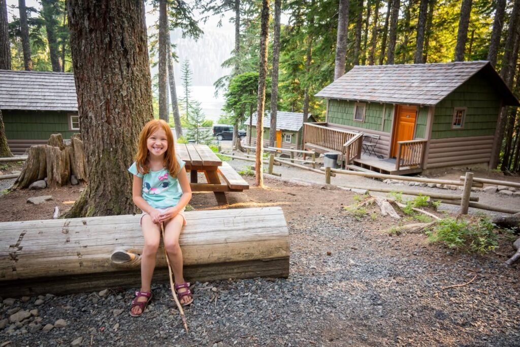 A person with red hair sits on a log bench in front of rustic wooden cabins surrounded by tall trees and forest scenery.