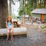 The Eclipse A person with red hair sits on a log bench in front of rustic wooden cabins surrounded by tall trees and forest scenery.