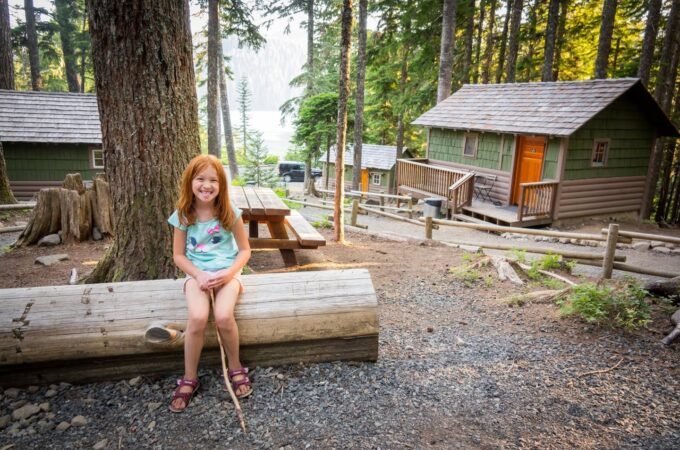 A person with red hair sits on a log bench in front of rustic wooden cabins surrounded by tall trees and forest scenery.