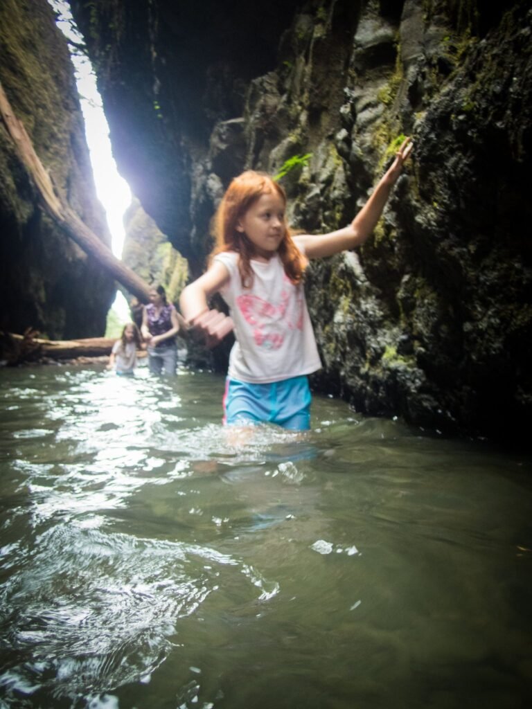 A traveler wades through a narrow gorge with towering rock walls beneath a sliver of sky. The calm water reflects the rugged landscape.