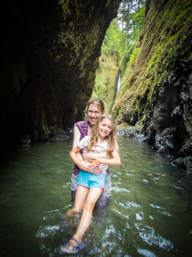 Two visitors enjoy a refreshing dip in a mossy natural pool within a lush Pacific Northwest canyon.