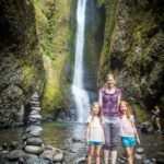 A woman and two young girls pose at the base of a tall, flowing waterfall surrounded by moss-covered rocks in the Pacific Northwest.