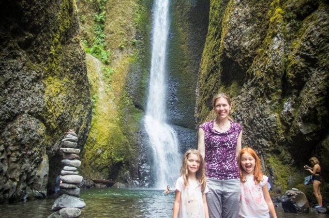 A woman and two young girls pose at the base of a tall, flowing waterfall surrounded by moss-covered rocks in the Pacific Northwest.