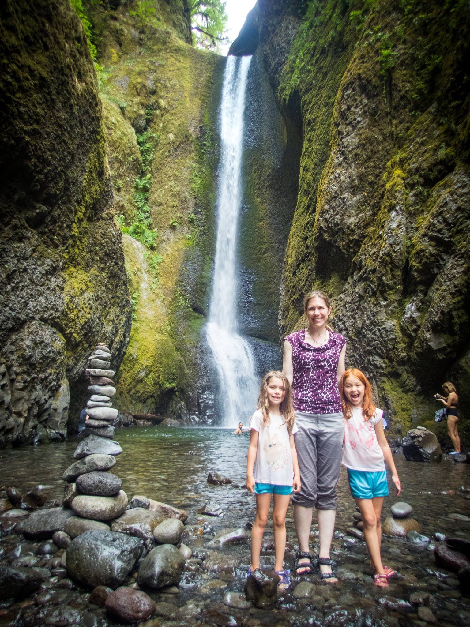 Oneonta Gorge A woman and two young girls pose at the base of a tall, flowing waterfall surrounded by moss-covered rocks in the Pacific Northwest.