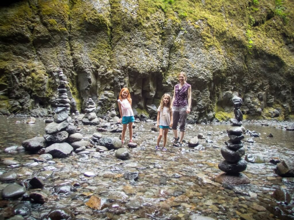 People standing in a shallow creek with mossy rock walls and stacked stone cairns.