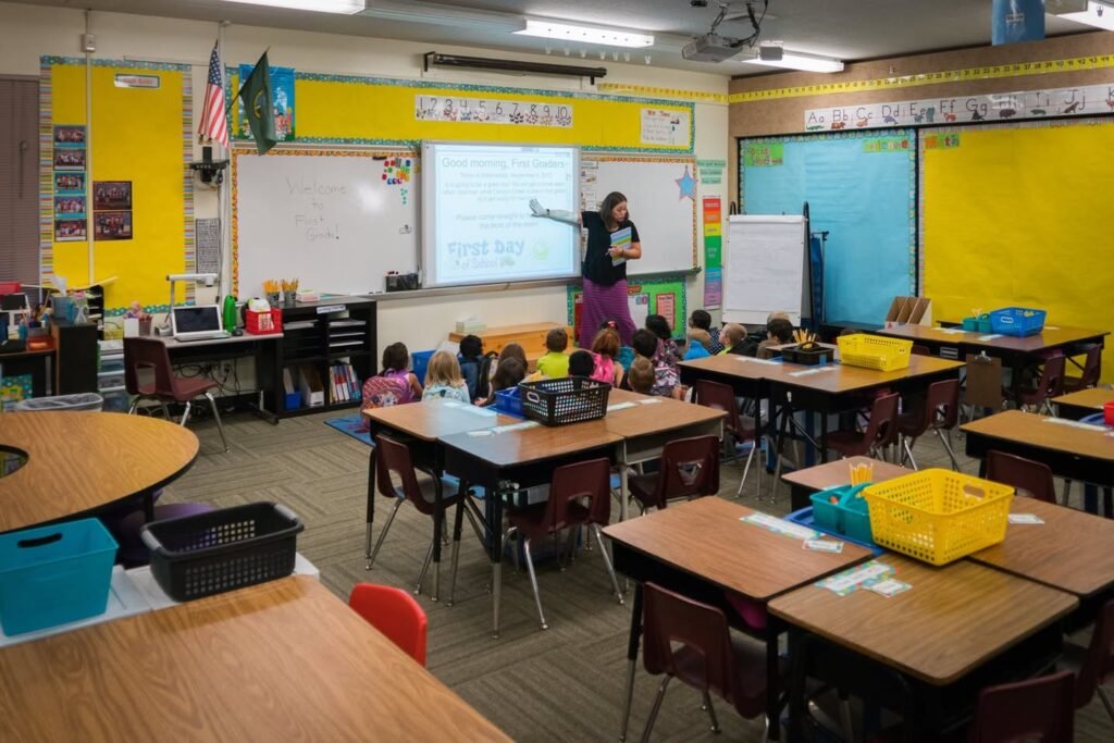 A teacher leads a lesson in a colorful classroom with children seated at tables.