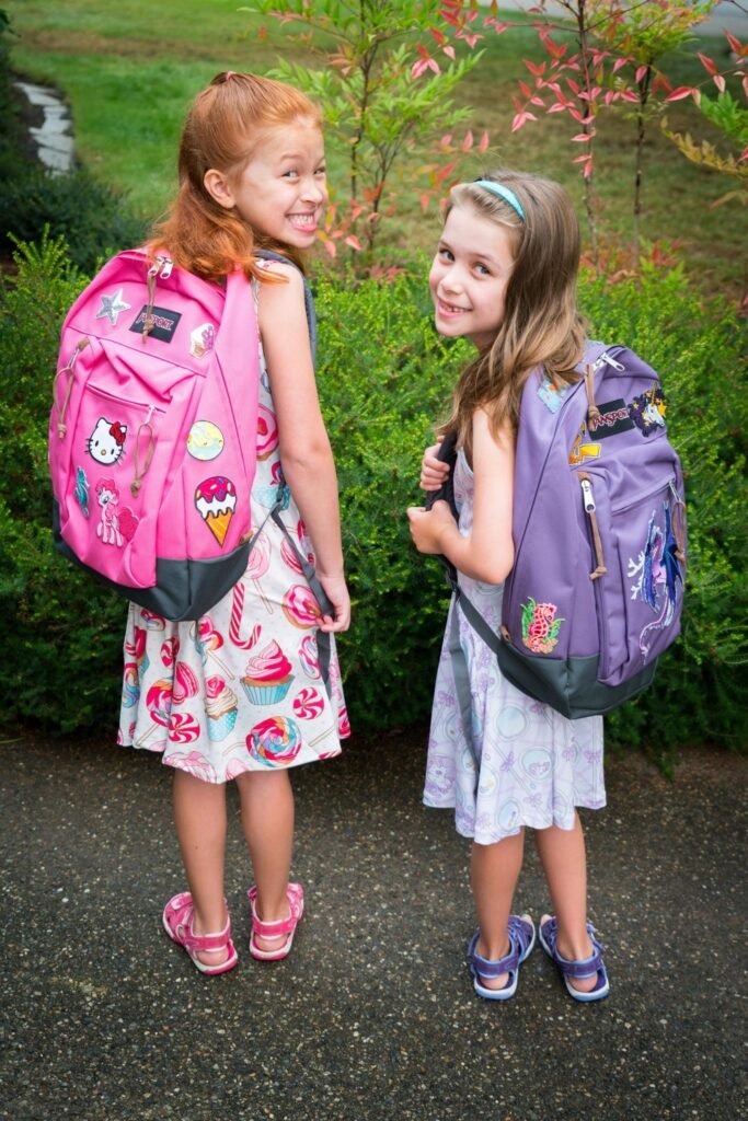 Two children show off their new pink and purple backpacks. They are standing outdoors on a paved path with greenery in the background.