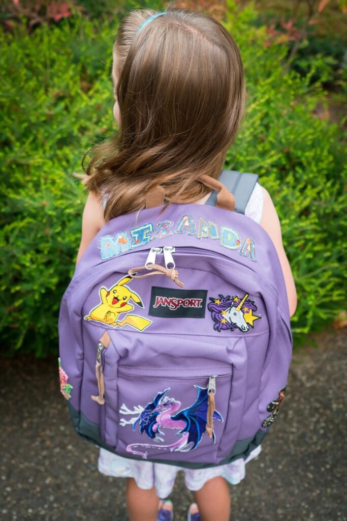 A child wears a purple school backpack featuring My Little Pony and Pokémon designs against a green garden background.