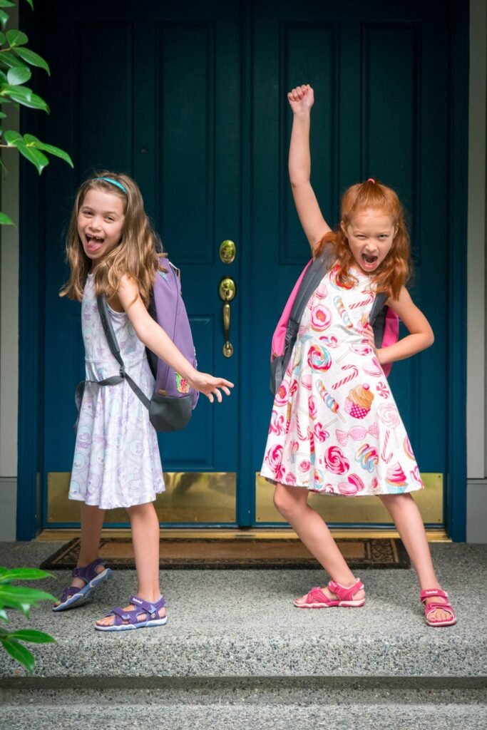 Two happy children wearing backpacks dance in front of a teal door.