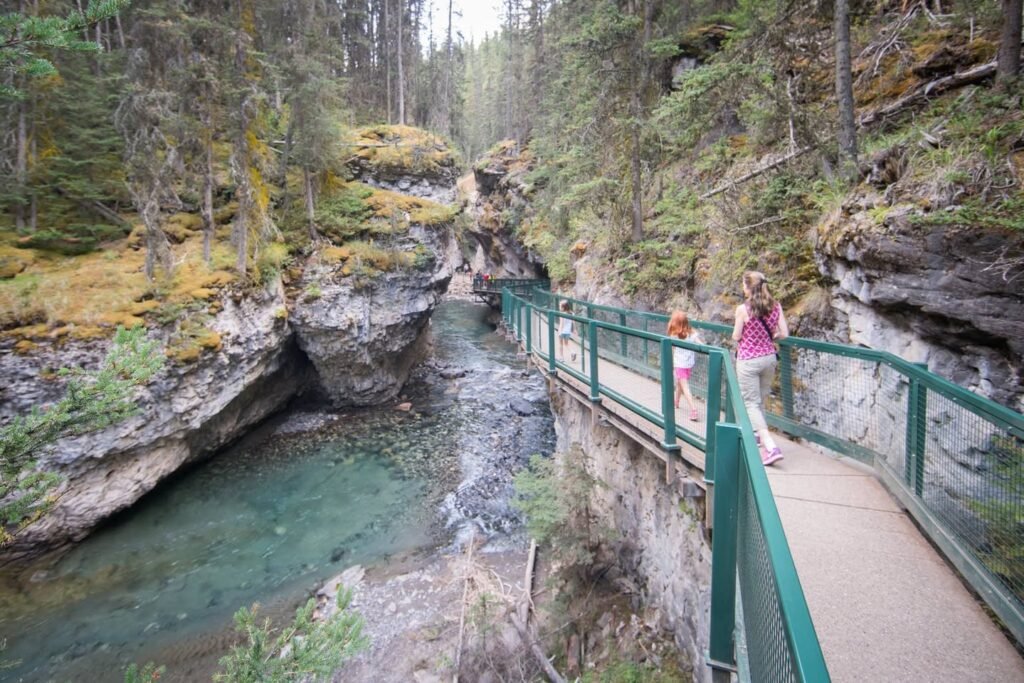 A wooden boardwalk with green railings bridges a narrow canyon with clear water. The path is surrounded by rocky terrain and evergreen trees in the Pacific Northwest.