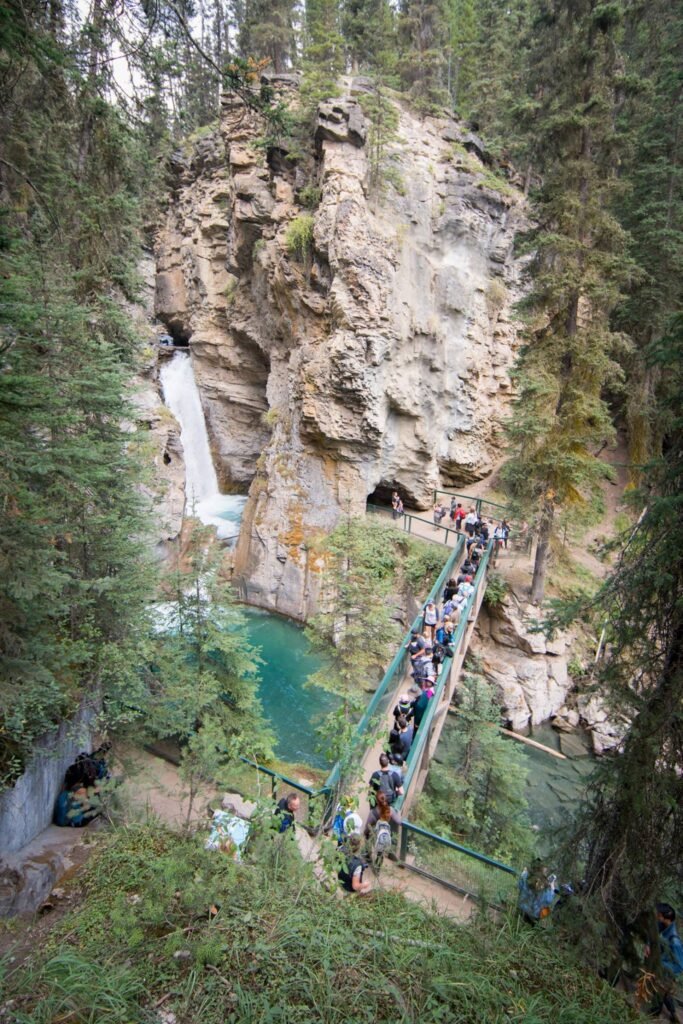 Tourists walk along a scenic pathway through a rugged rock canyon with a waterfall and emerald pool. Lush green trees frame the steep cliffs and natural landscape.