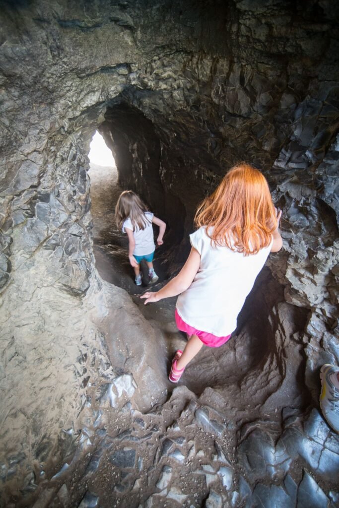 Two children explore a rugged sea cave. Natural light streams through an opening in the rocky wall.