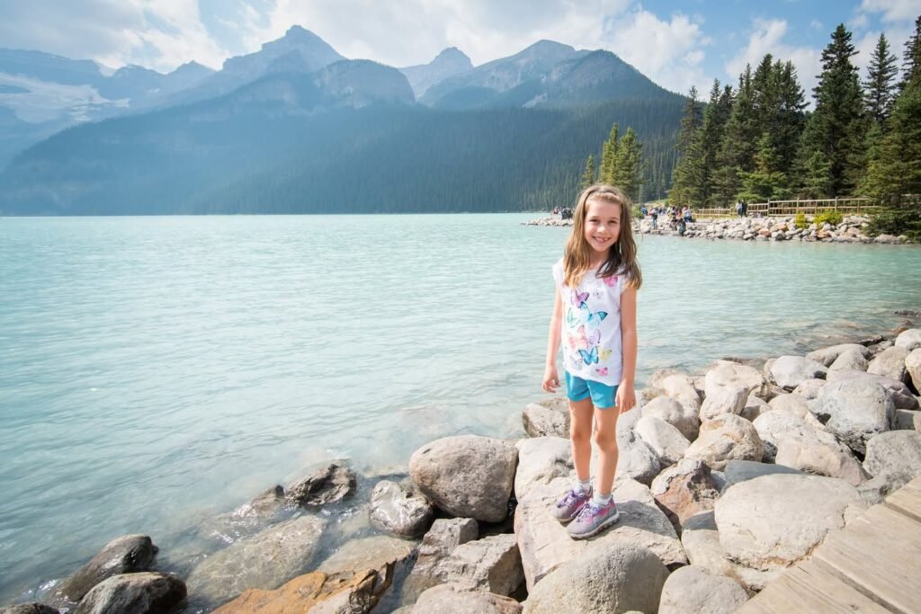 A scenic view of a rocky lakeshore with evergreen trees and mountain peaks under a clear sky.