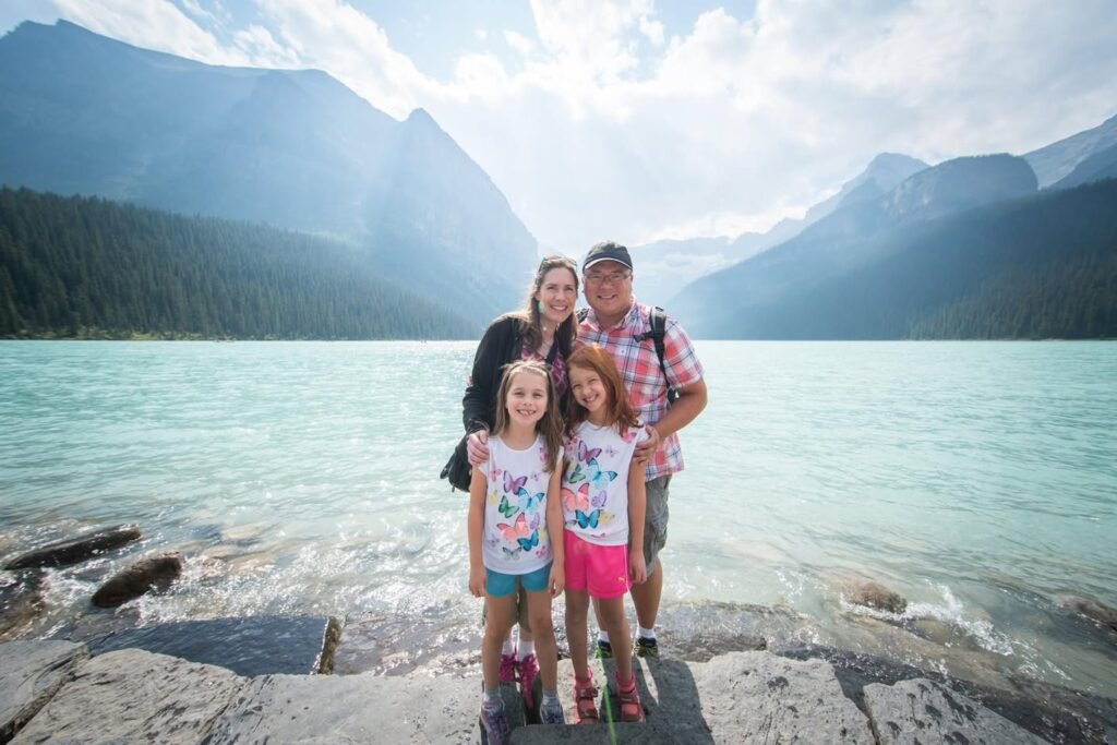 A family poses on a rocky shoreline with a turquoise lake and towering mountains in the background. The distinctive teal water and rugged landscape are characteristic of Moraine Lake in Banff National Park.