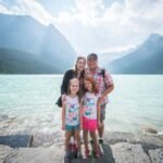 Johnston Canyon Hike A family poses on a rocky shoreline with a turquoise lake and towering mountains in the background. The distinctive teal water and rugged landscape are characteristic of Moraine Lake in Banff National Park.