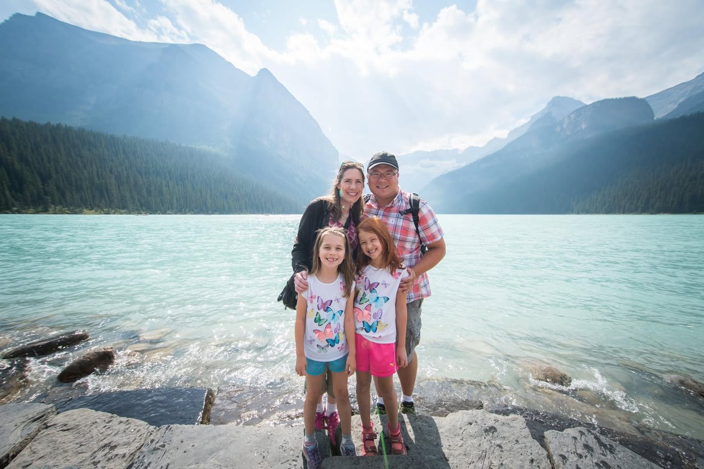 A family poses on a rocky shoreline with a turquoise lake and towering mountains in the background. The distinctive teal water and rugged landscape are characteristic of Moraine Lake in Banff National Park.