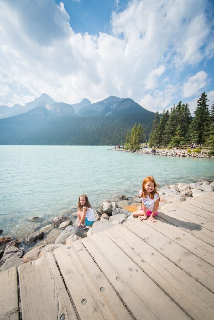 Two children sit on a rustic wooden dock by a clear, calm lake under a partly cloudy sky. A misty mountain range rises in the distant background.