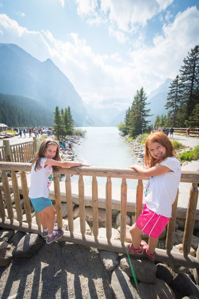 Young girls lean on a wooden fence overlooking a serene mountain lake in the Canadian Rockies.