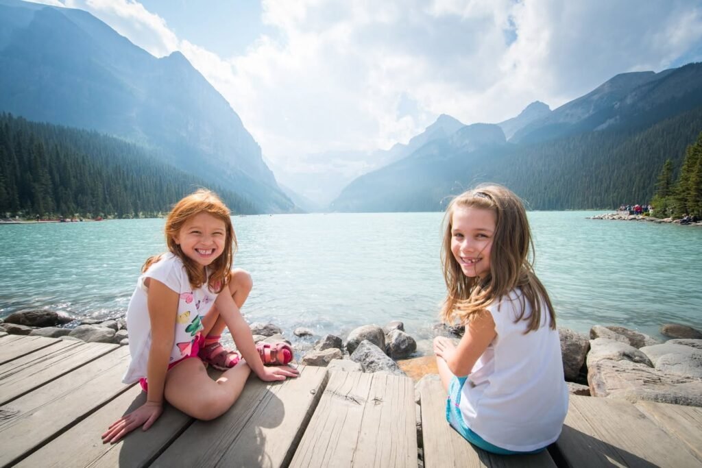Two kids sitting on a wooden dock overlooking a turquoise lake surrounded by mountains.