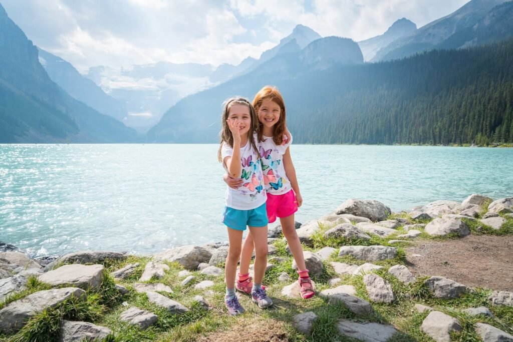 Children standing on a mossy rock shore beside a turquoise mountain lake. The background features dense evergreen forests and towering peaks.