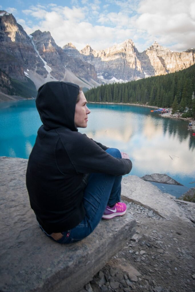 A traveler in a black hoodie sits on a rock overlooking a stunning turquoise alpine lake framed by rugged mountain peaks.