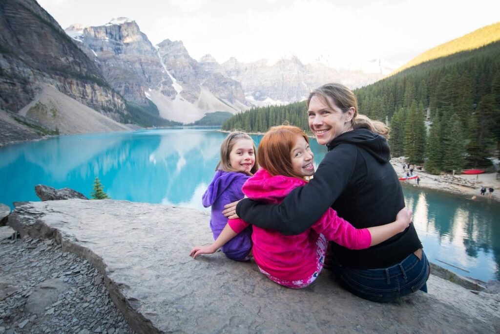 A family shares a warm embrace while sitting by a stunning turquoise lake surrounded by rugged mountain peaks and evergreen forests.