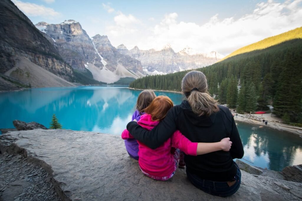 View from a rocky outcrop overlooking Moraine Lake, Alberta, with snow-capped mountains and evergreen forests. Individuals are seated together, gazing at the stunning turquoise water.