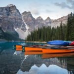 Canoes on a mountain lake reflect rocky peaks, evergreen lakeside, and colorful canoes.