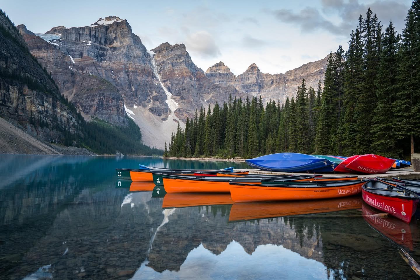 Moraine Lake Canoes on a mountain lake reflect rocky peaks, evergreen lakeside, and colorful canoes.