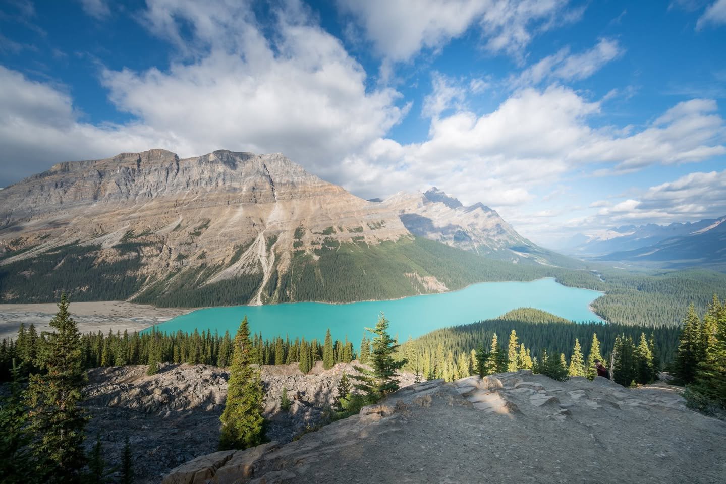 A panoramic view of a turquoise alpine lake surrounded by rugged mountains and pine trees under a blue sky.