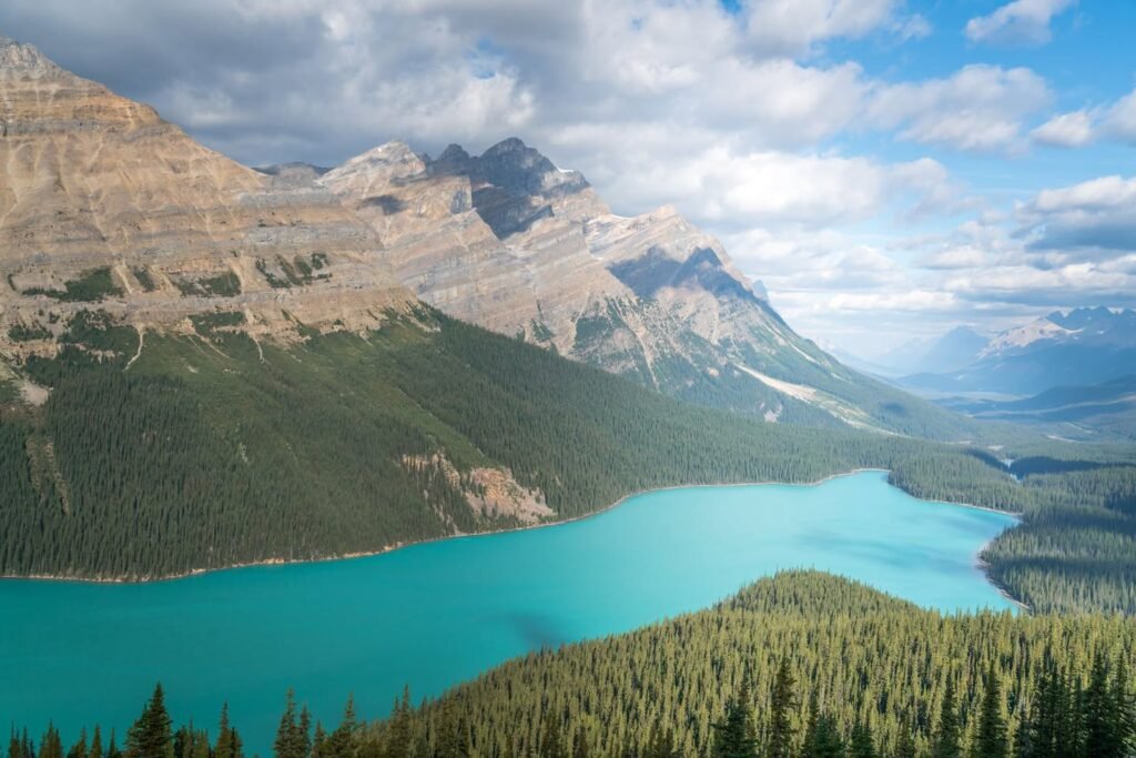 A scenic view of a turquoise mountain lake surrounded by dense evergreen forests and towering rocky peaks under a cloudy sky.