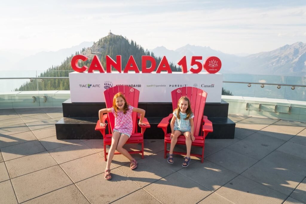 Two children sit on red chairs in front of a Canada 150 sign with a mountainous background.