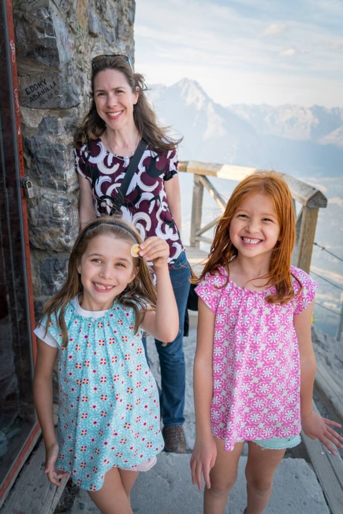 A group of four people, including two young girls and an adult, smiling at a scenic overlook with mountain views.