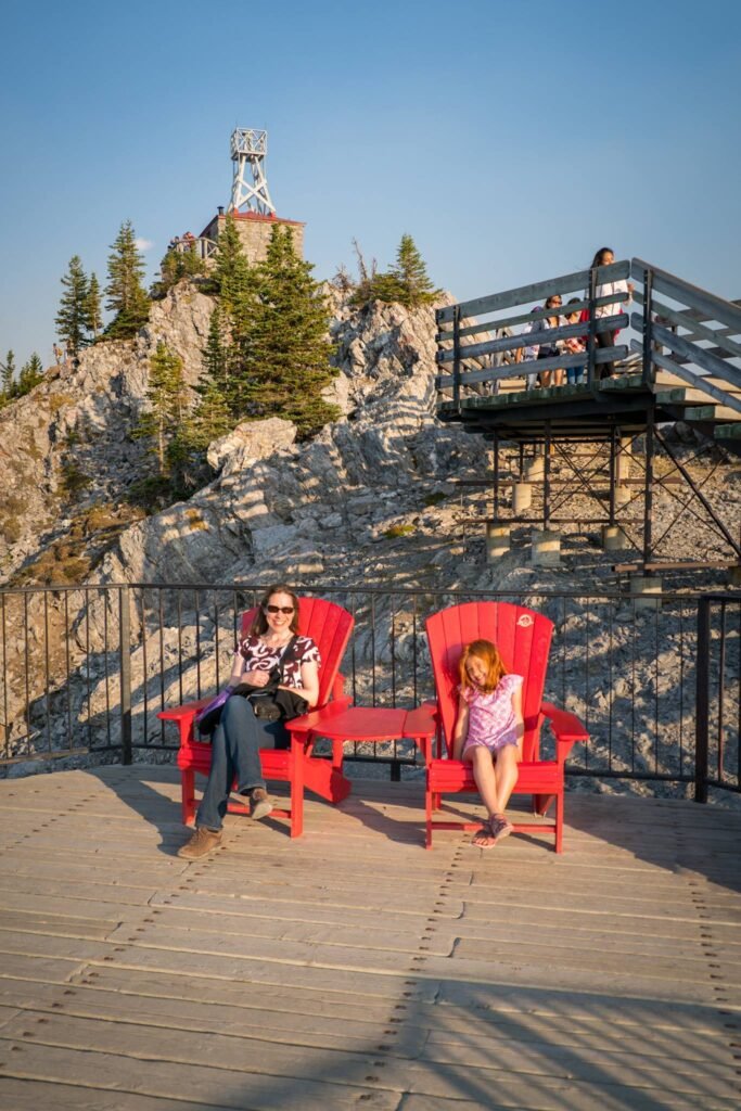 A person and a child relaxing in red Adirondack chairs at an outdoor mountain viewpoint. In the background, a fire lookout tower is visible atop a rocky hill covered with evergreen trees.