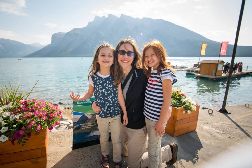 A family smiles together on a sunny day at a lakeside dock with colorful flowers and mountains in the background.
