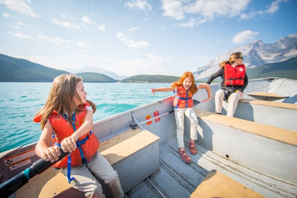 People enjoying a boat ride on a turquoise mountain lake. The scene features snow-capped peaks and a bright blue sky.