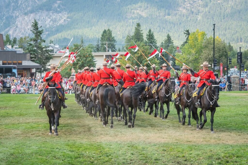 Mounted officers in red uniforms parade on horseback through a mountain town during a ceremonial event.