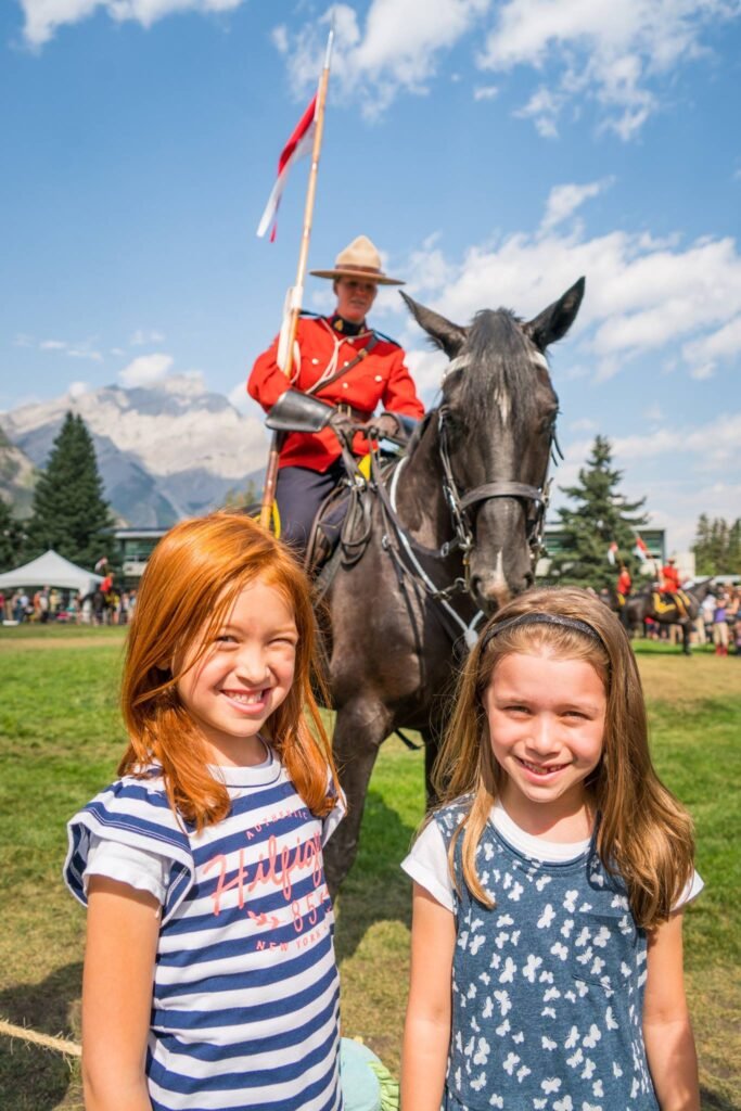 Two girls posing in front of a mounted Royal Canadian Mounted Police officer with a waving flag at an outdoor event. Snow-capped mountains and evergreen trees provide a scenic background.