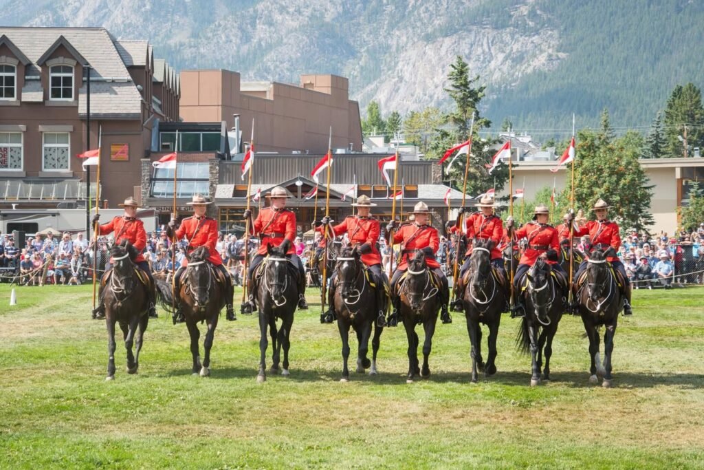 The Royal Canadian Mounted Police march in ceremonial attire. A group of officers on horseback is seen in a formation on a grassy field with a historic building and mountains in the background.