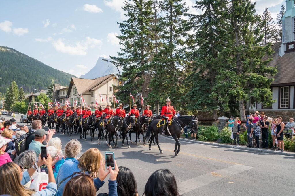 Mounted police officers in red tunics march through a scenic mountain town as spectators watch from the roadside. Fir trees and Alpine buildings frame the procession under a sunny sky.