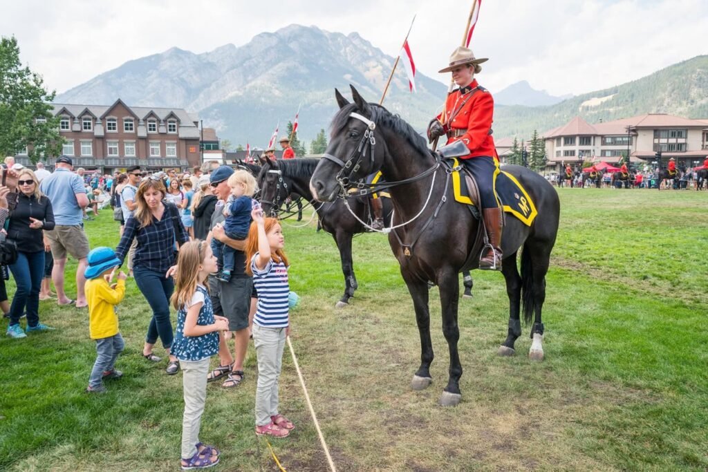 A line of dark horses with uniformed riders marches on a grassy field during a local festival. Families gather along the perimeter under a cloudy sky, set against a backdrop of mountains and a red-roofed lodge.