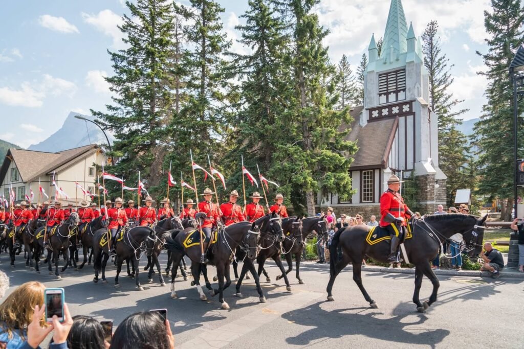Uniformed riders on horseback parade past a church tower with tall evergreens and a mountain in the distance.