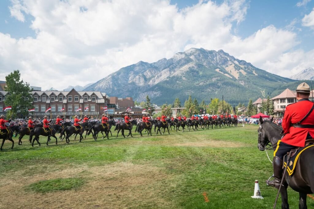 A line of Red Coat Mounties rides on a grassy field set against a large mountain range and historic buildings.
