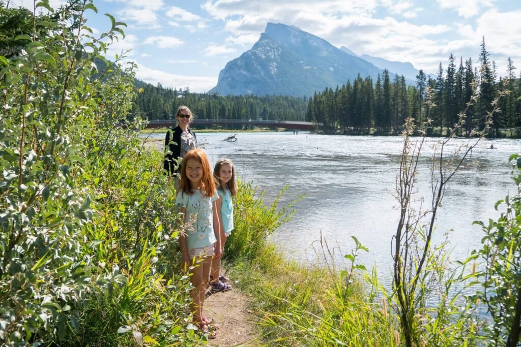 A family walks along a lakeside trail surrounded by lush vegetation with a prominent mountain peak in the background.